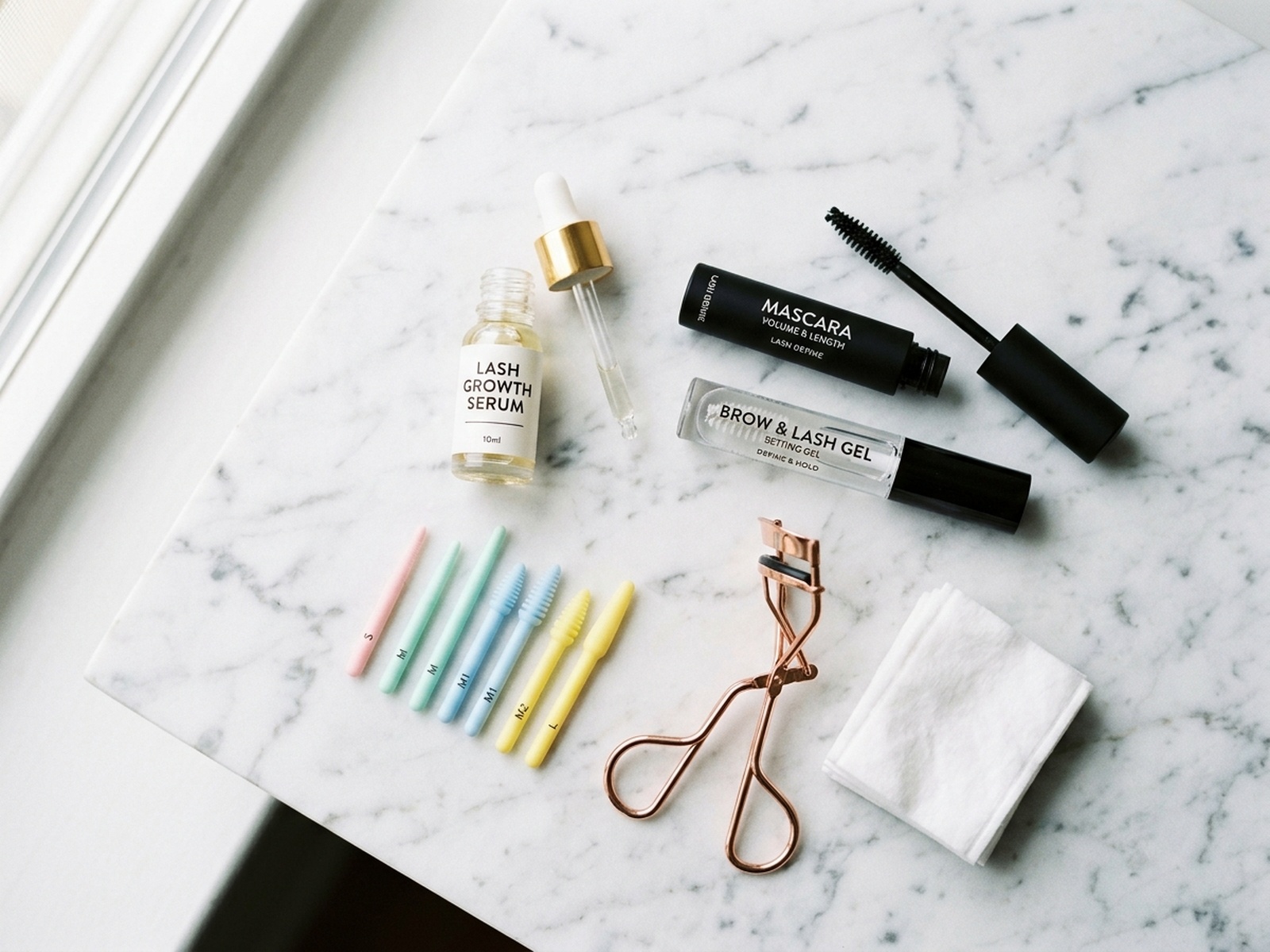 Productos de cuidado de pestanas: suero, rizador, mascara y varillas de lifting en marmol blanco